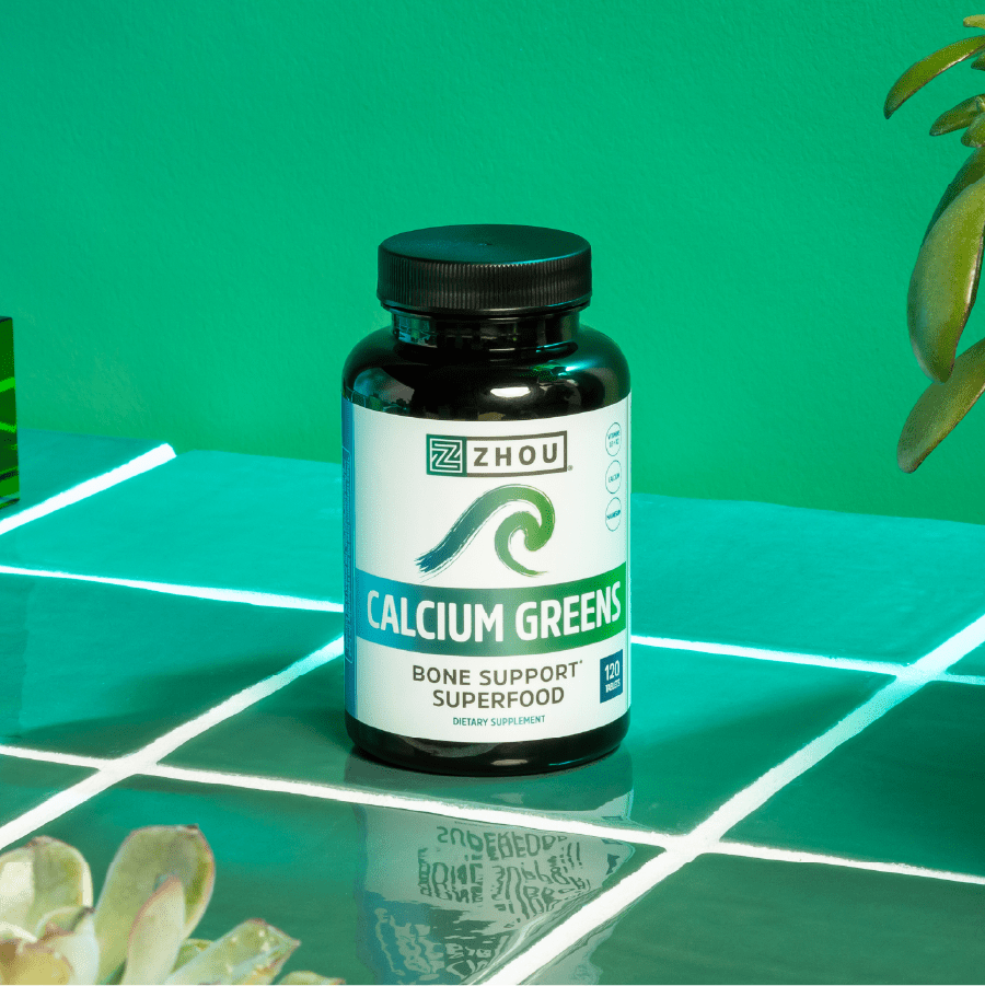 A bottle of Zhou Nutrition Calcium Greens supplement sits on tiles with green plants in the background.